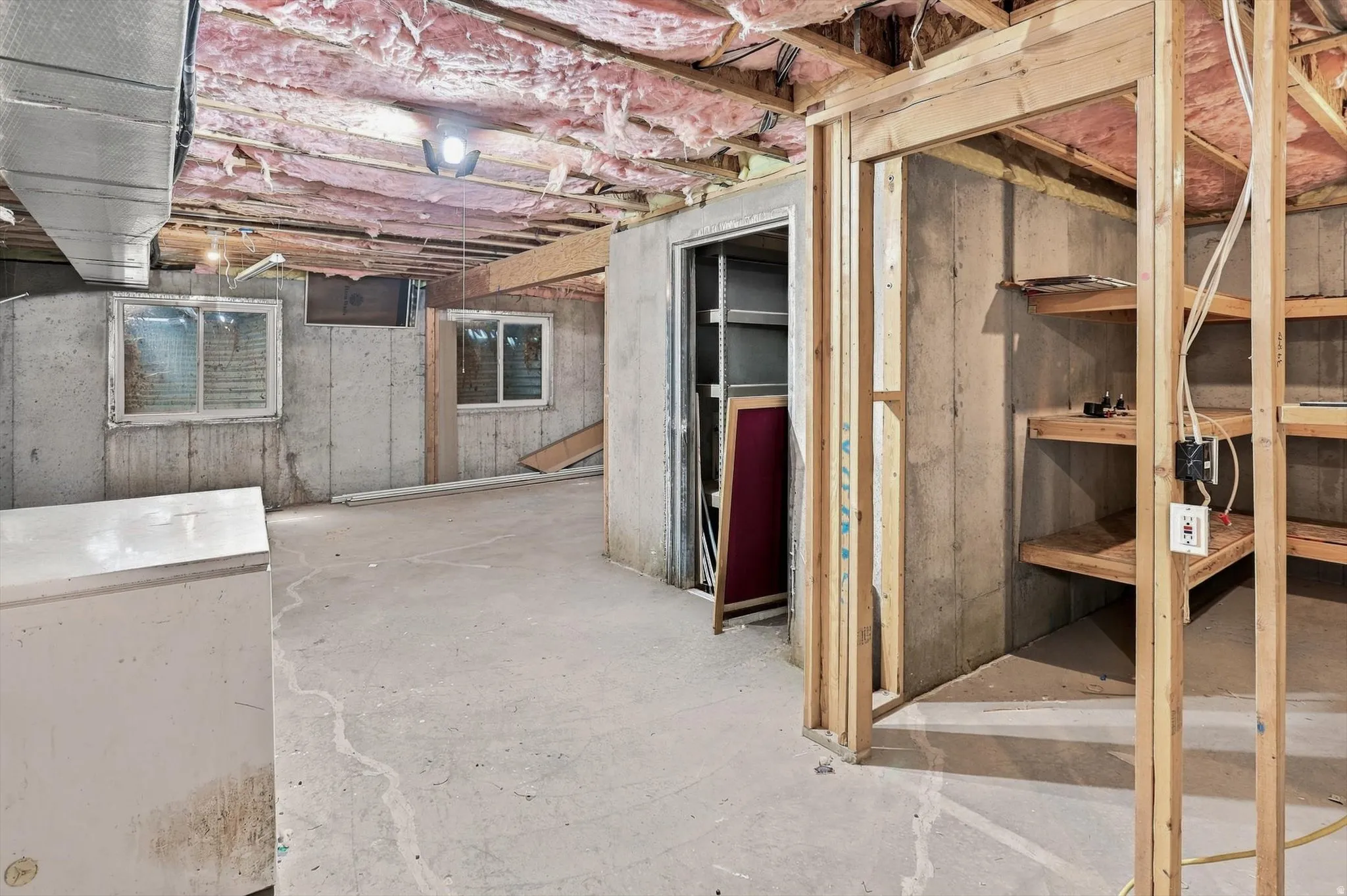 Unfinished basement with exposed ceiling joists and insulation, concrete flooring, and built-in wood shelving