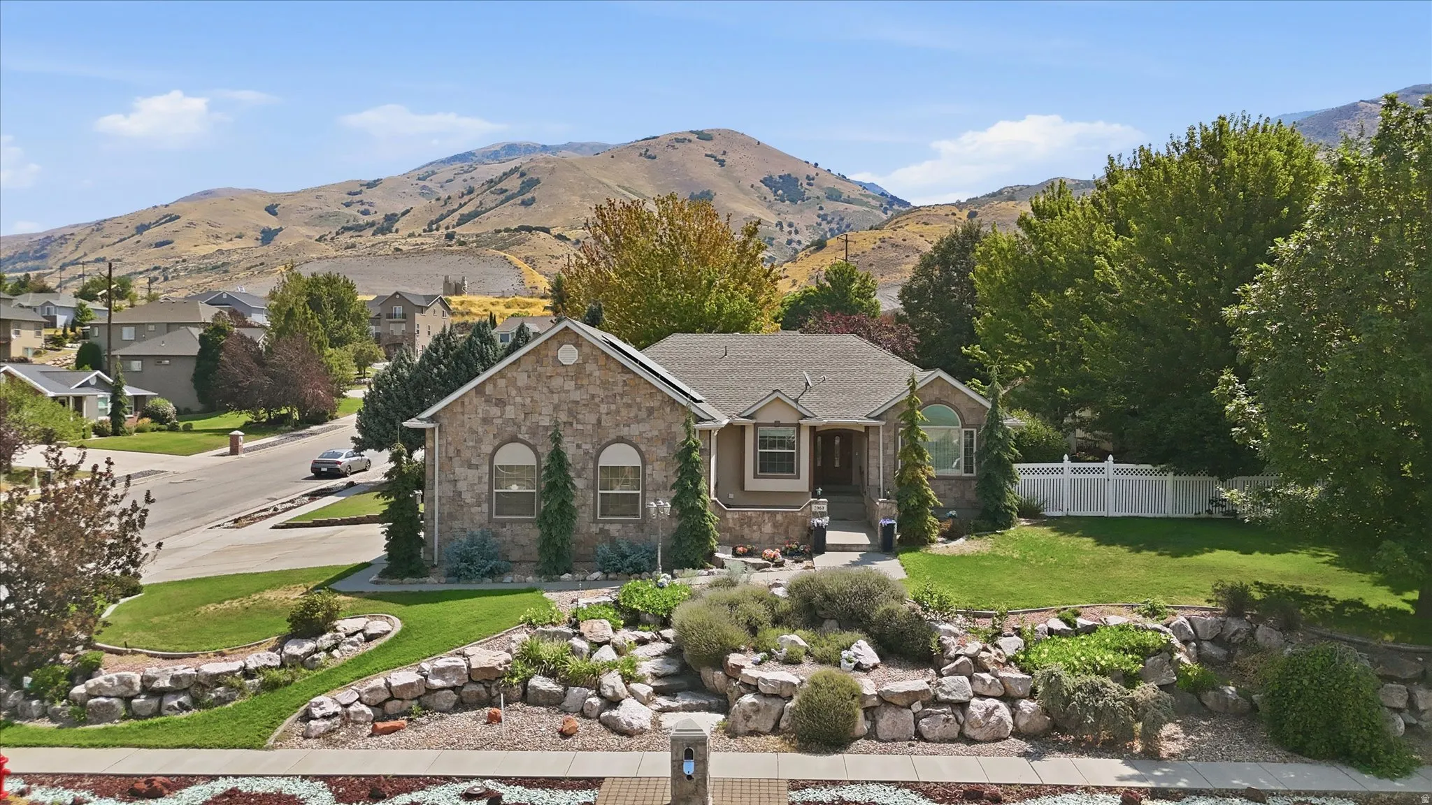 Stone exterior with a gabled roofline, mature trees, manicured lawn, white privacy fencing, and terraced landscaping