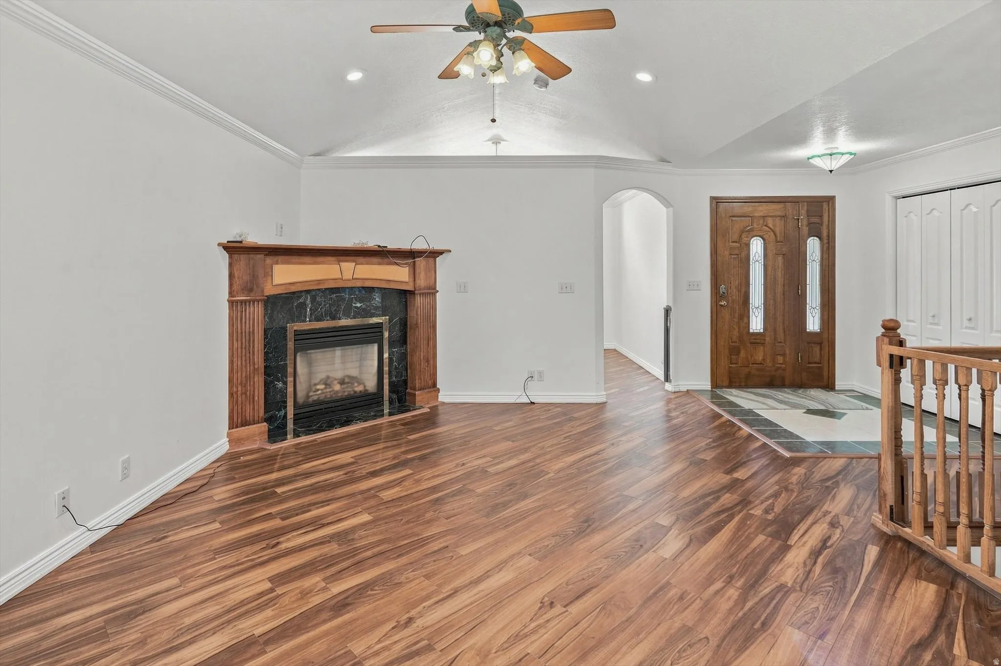 Vaulted ceiling with recessed lighting and ceiling fan, wood-finish flooring, wood fireplace surround with dark stone, detailed wooden entry door, and architectural archway