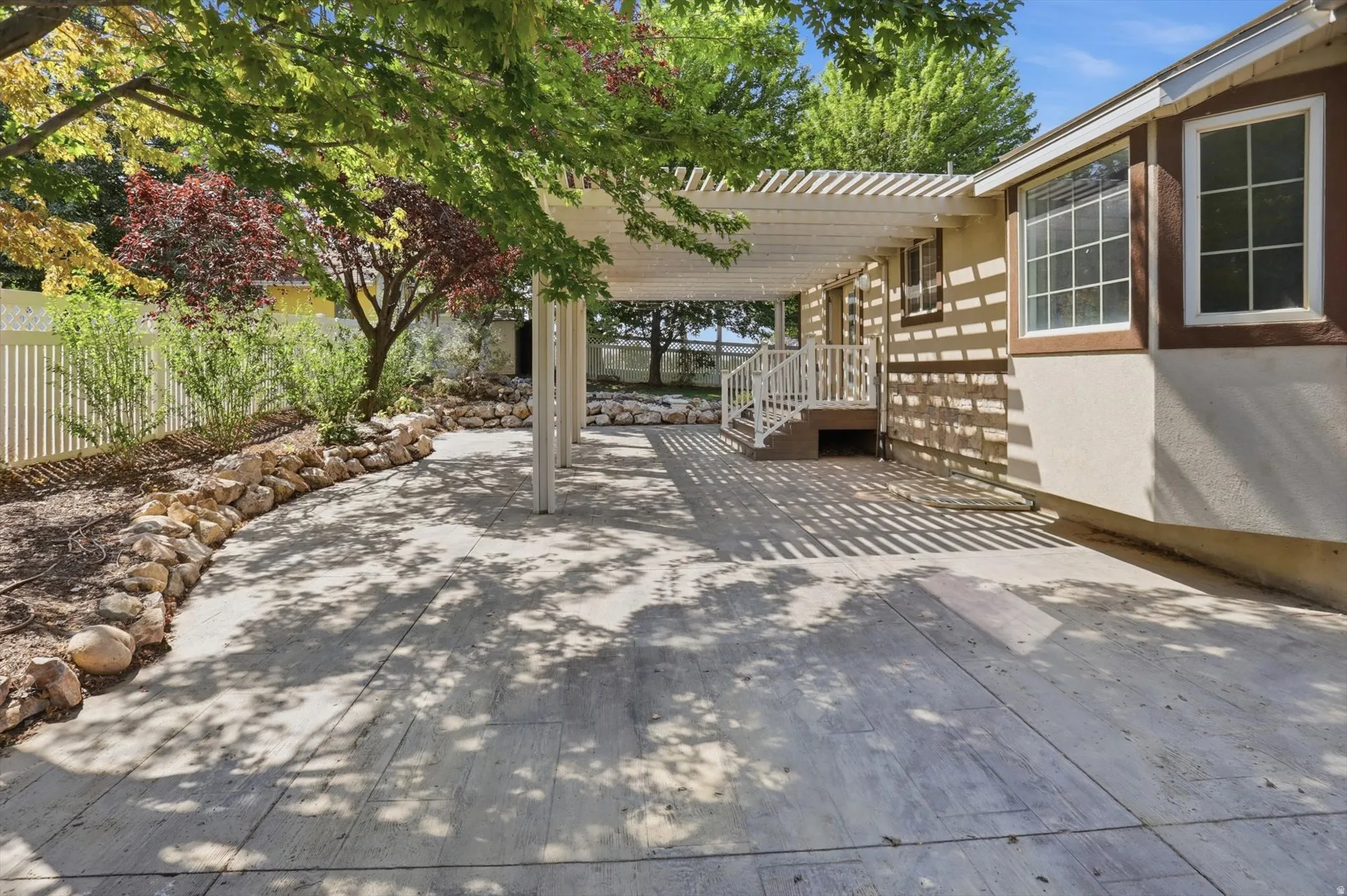 Expansive concrete patio with a white pergola, stone-lined garden beds, and a white picket fence