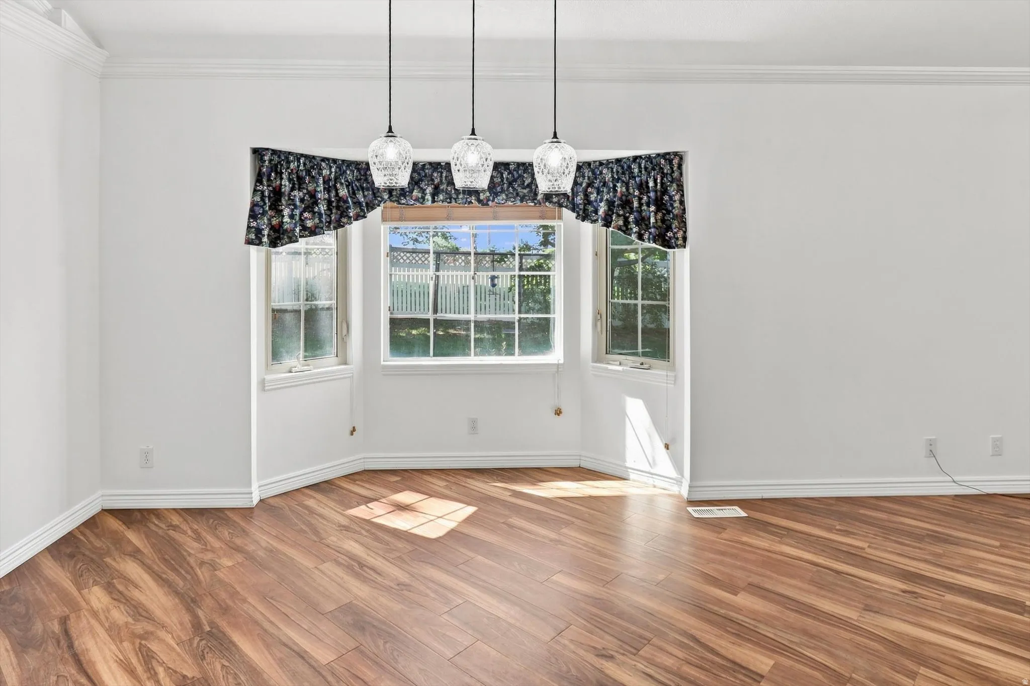 Bay window with multiple panes, wood-finish flooring, three pendant light fixtures, crown molding, and white wall finishes