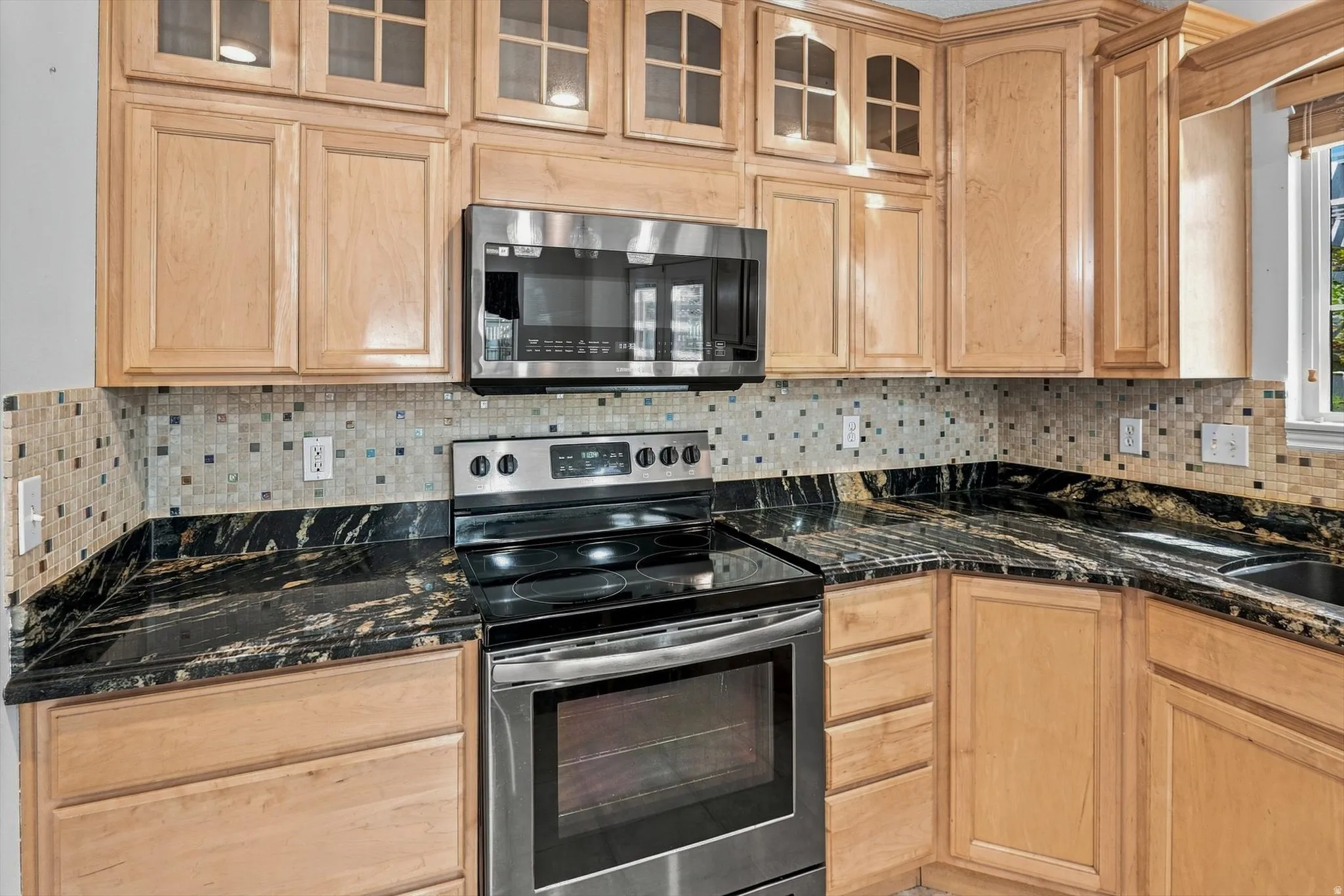 Light wood-finish cabinetry with glass upper door fronts, dark countertops, a mosaic tile backsplash, and stainless steel appliances