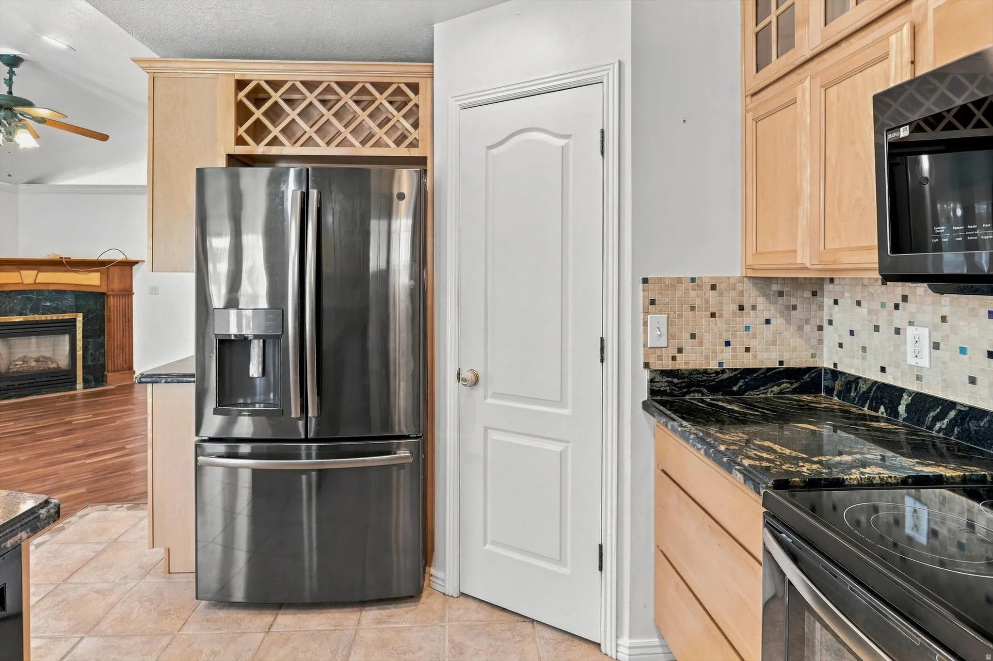 Stainless steel refrigerator, built-in wine rack, black granite countertops, mosaic tile backsplash, and light wood cabinetry