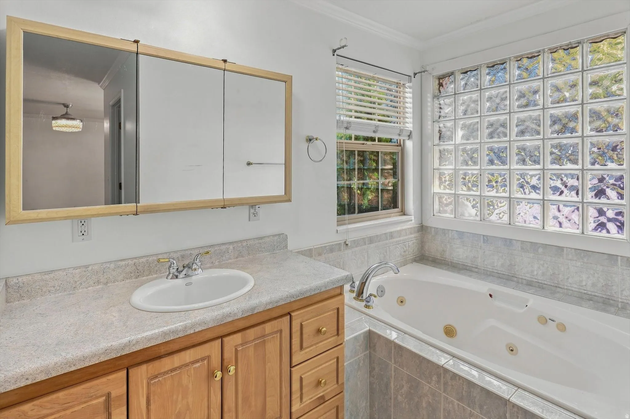 Bathroom featuring a jetted tub, glass block window, wood-finish vanity with a laminate countertop, and a mirrored medicine cabinet