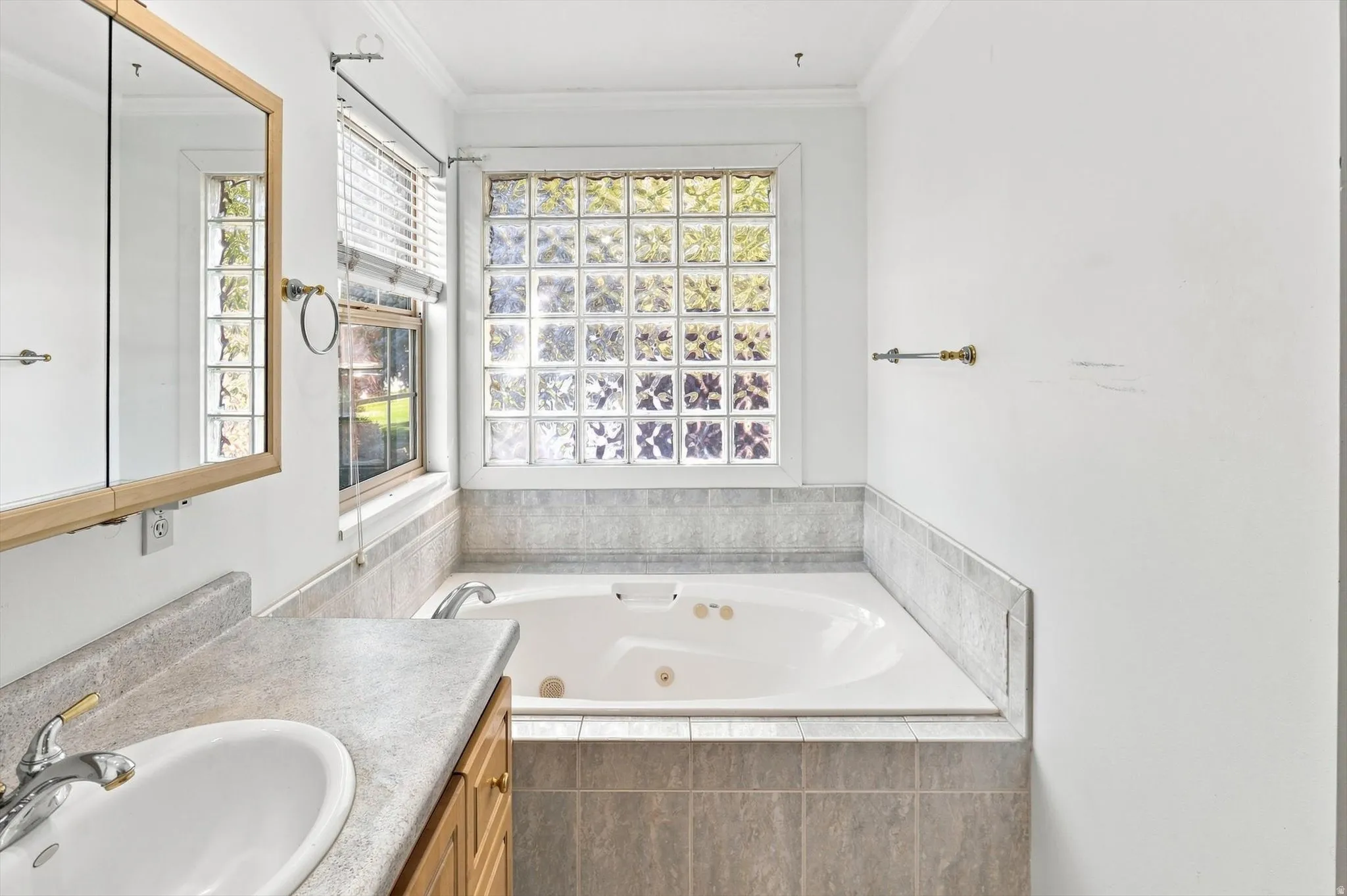Soaking tub with tiled surround, glass block window, wood-finish vanity with countertop, and integrated sink