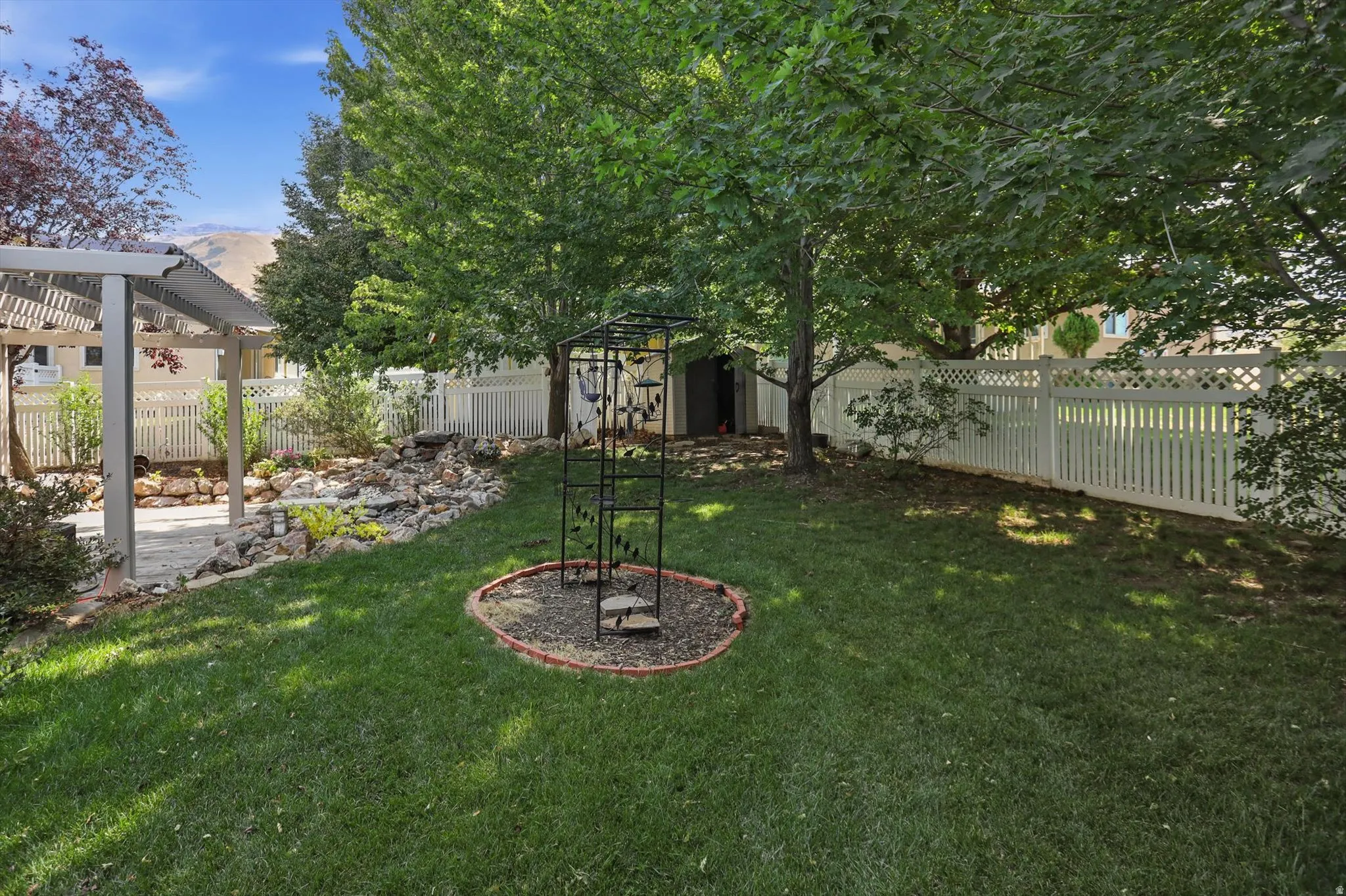 Expansive backyard with a manicured lawn, established trees, white picket fencing, and a built-in pergola over a paved patio
