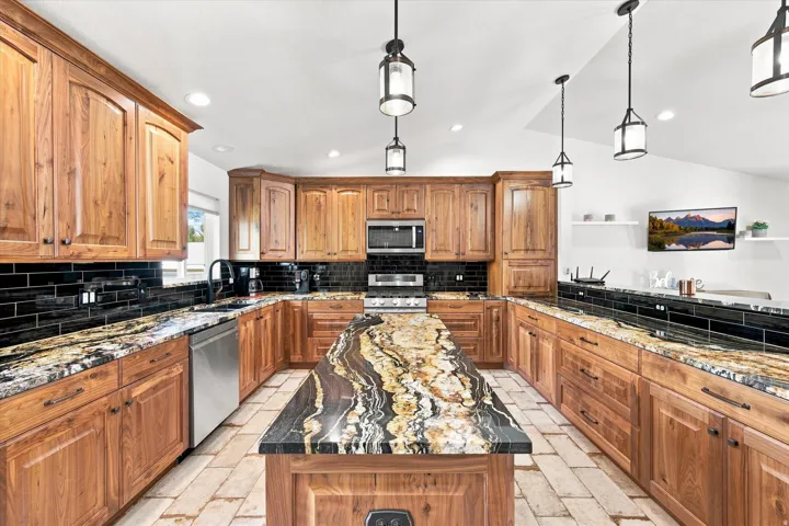 Wood cabinetry, black subway tile backsplash, stainless steel appliances, variegated countertops, and pendant lighting