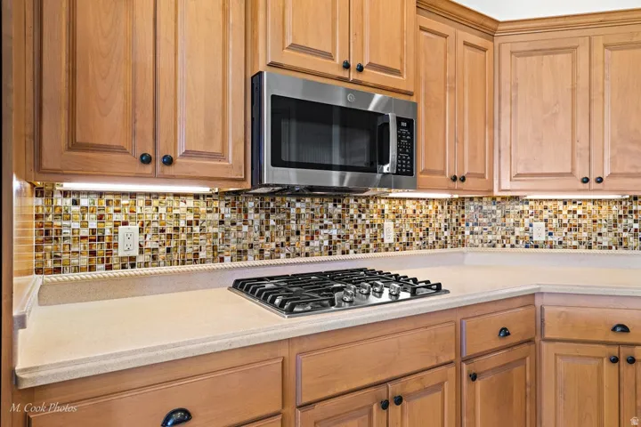 Light wood-finish cabinetry with black hardware, stainless steel built-in microwave, tiled backsplash, gas cooktop, and light-toned countertops