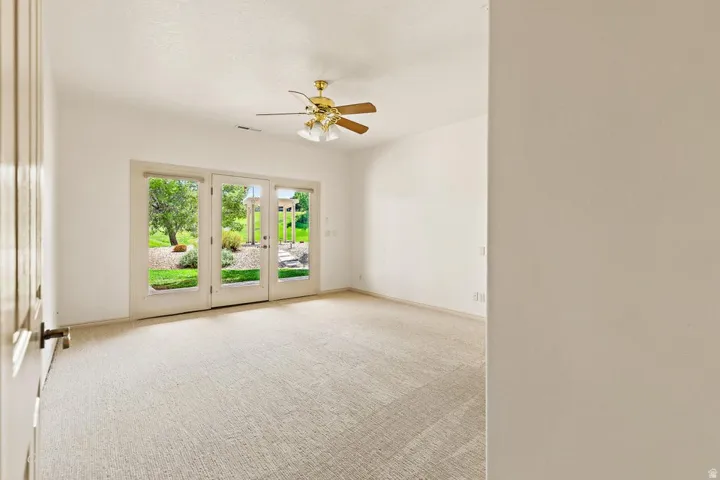 White walls, light-toned carpeting, ceiling fan with light fixture, and French doors leading to a landscaped outdoor area
