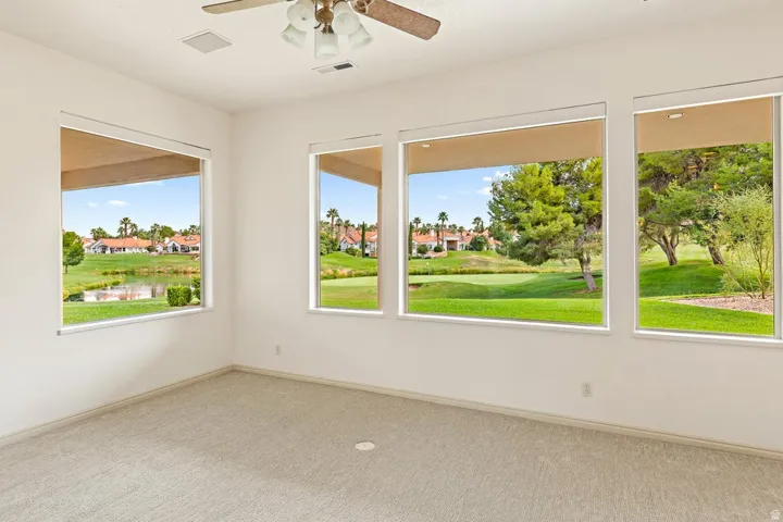 Expansive windows featuring views of a golf course and water feature, light-toned carpeting, and a ceiling fan with integrated lighting
