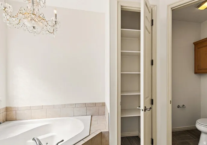 Jetted soaking tub with tiled surround, elegant chandelier, recessed shelving, and a wood-finish floor