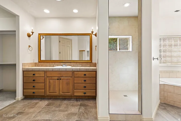Spa-inspired primary bathroom featuring a custom wood vanity with multiple drawers and cabinet storage, framed mirror with warm wood trim, dual pendant lighting, porcelain tile flooring, a spacious walk-in shower with glass enclosure and subway tile surro