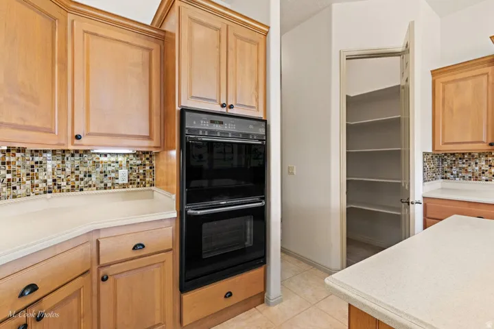 Light wood cabinetry, built-in double oven, mosaic tile backsplash, light-toned countertops, and walk-in pantry with shelving