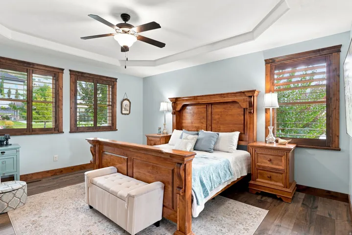 Main floor primary bedroom 1. Tray ceiling with a ceiling fan, wood-finish flooring, two windows with wood plantation shutters, and wood trim accents