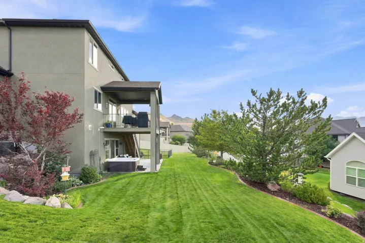 Expansive backyard featuring a manicured lawn, established landscaping, and a covered lower-level patio with an integrated hot tub