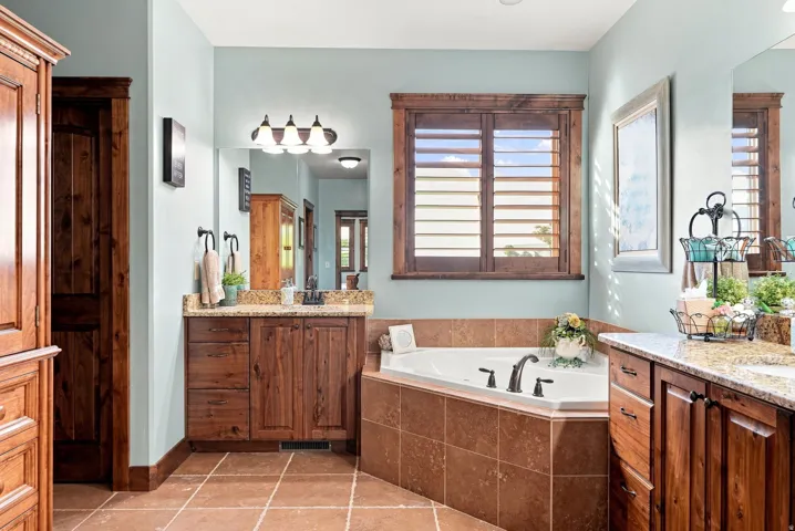 Corner soaking tub, dual wood-finish vanities with granite countertops, tile flooring, and wood plantation shutters