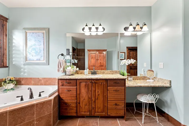 Primary Bathroom. Corner soaking tub with tiled surround, wood-finish vanity with integrated sink, two large mirrors, dual wall-mounted vanity lights, and a stone-look countertop