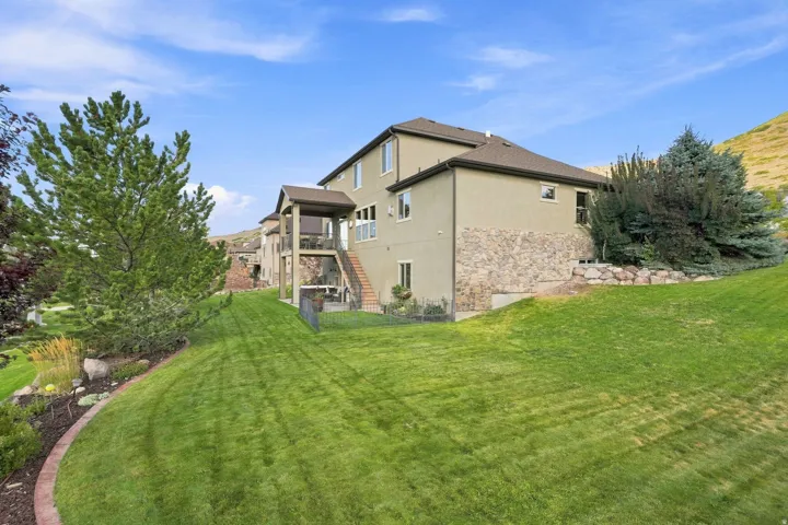 Expansive backyard featuring a lush lawn, established landscaping, stone veneer exterior accents, and a covered patio with direct stair access