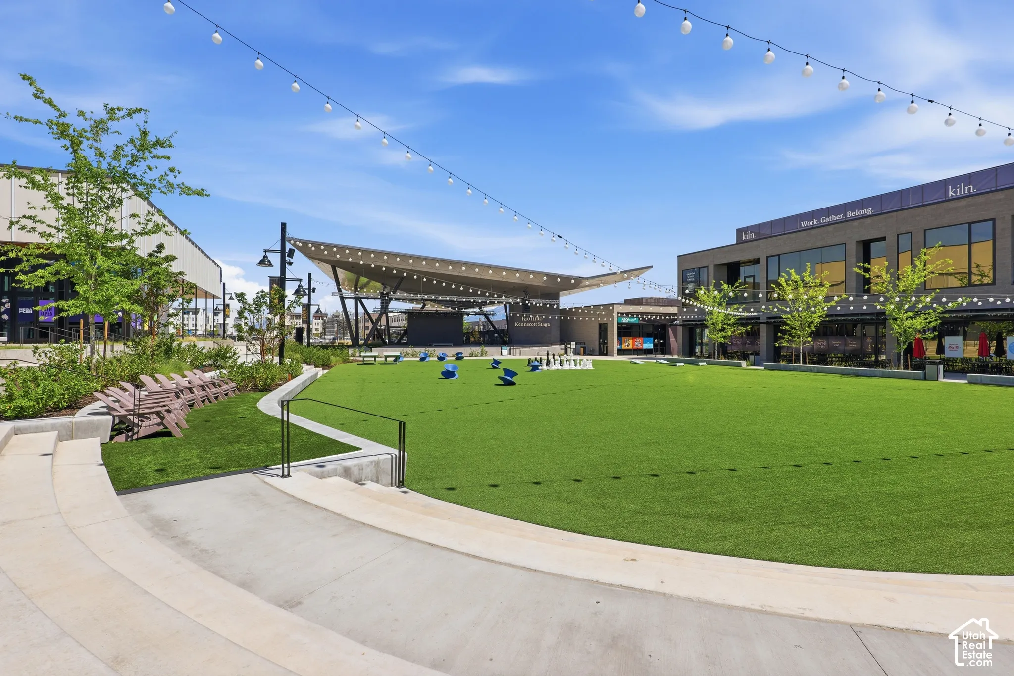 Expansive green space with concrete tiered seating, a covered outdoor stage, and string lighting