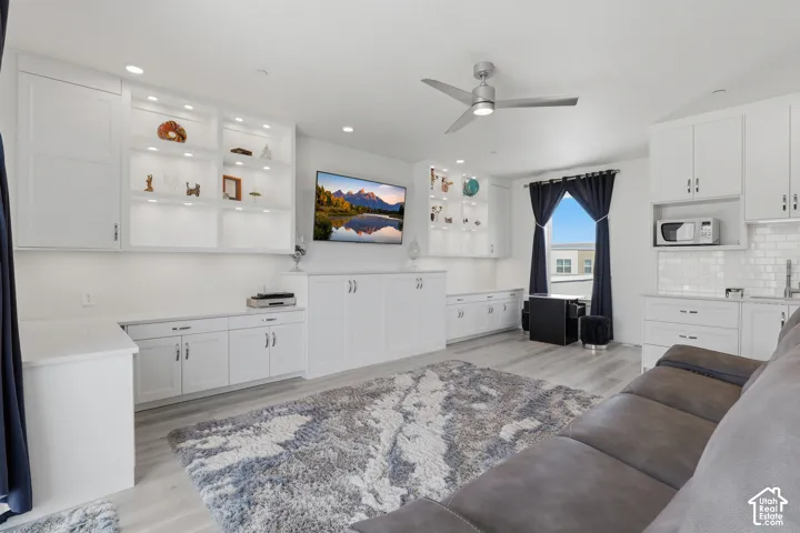 Built-in cabinetry with illuminated display shelving, contemporary ceiling fan, light wood-finish flooring, white subway tile backsplash, and a window with dark drapery