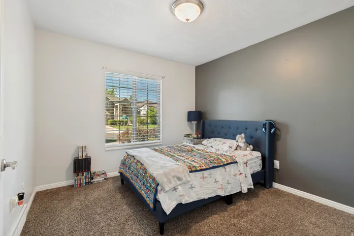 Spacious bedroom featuring a neutral color palette with an accent wall in soft gray, abundant natural light from a large window with blinds, neutral carpet flooring, and a ceiling-mounted light fixture