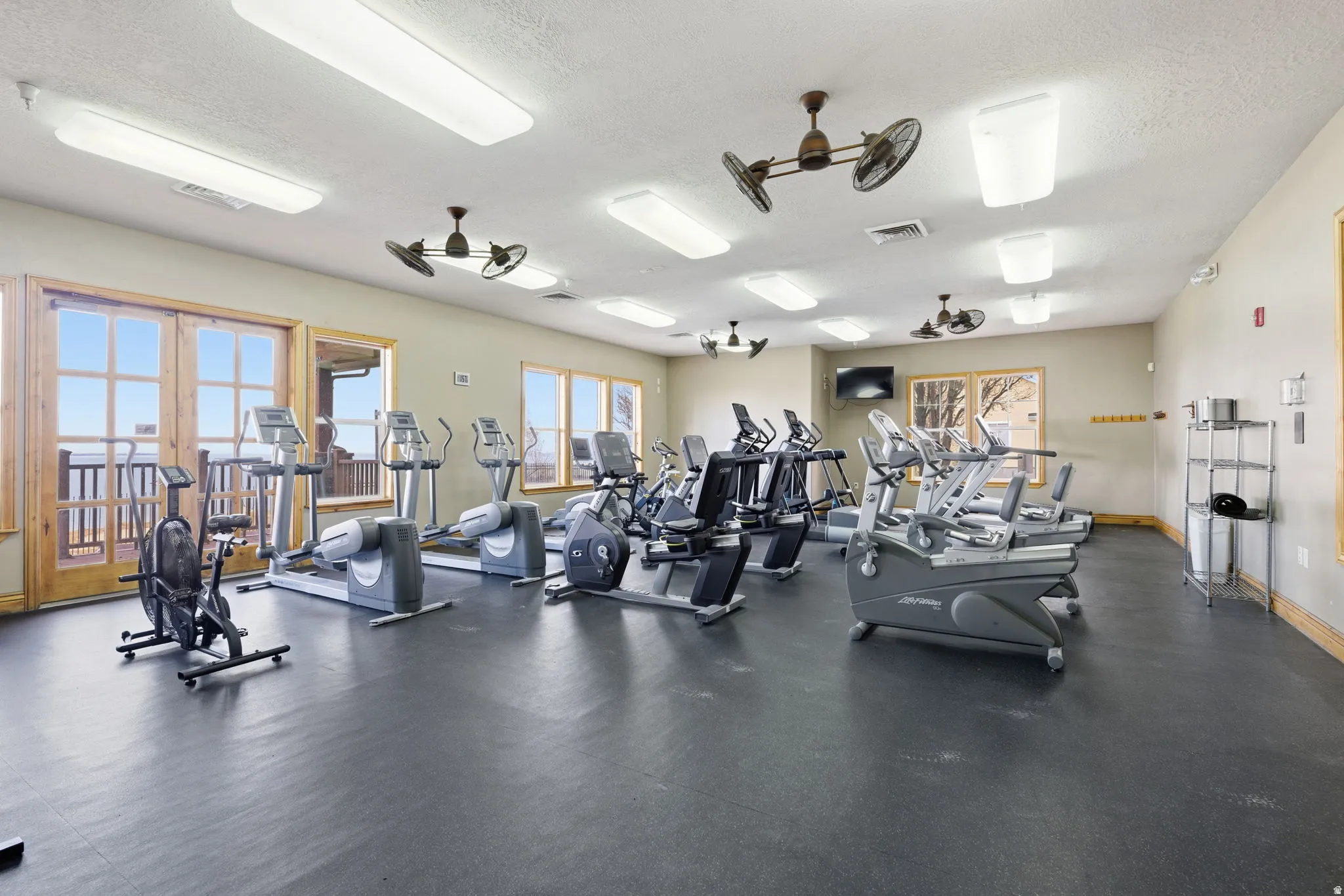 Fitness facility with rubber flooring, extensive cardio equipment, multiple windows, double doors, and ceiling-mounted fans