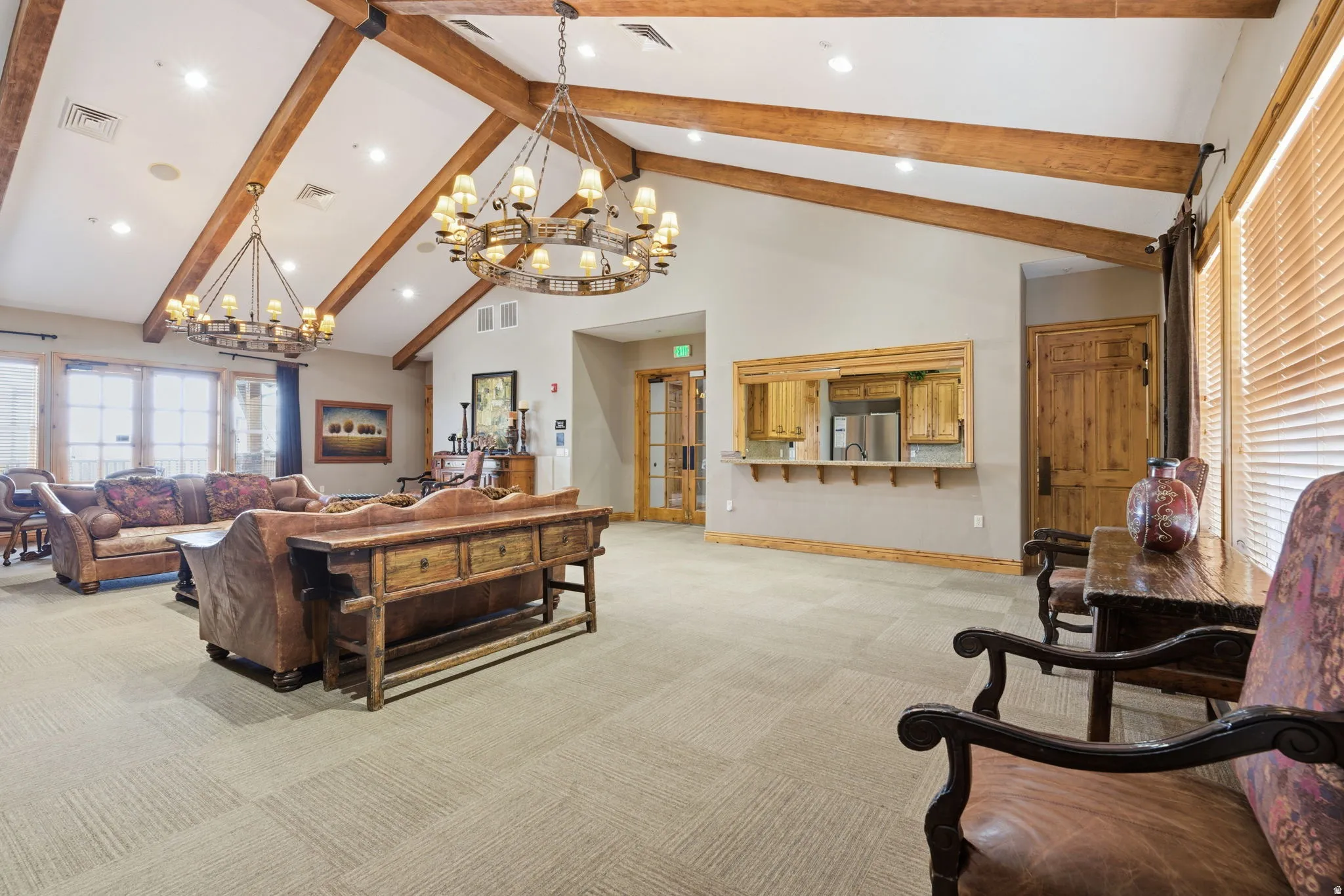 Vaulted ceiling with exposed wood beams, two large circular chandeliers, light-toned carpet flooring, and a built-in serving area with wood cabinetry