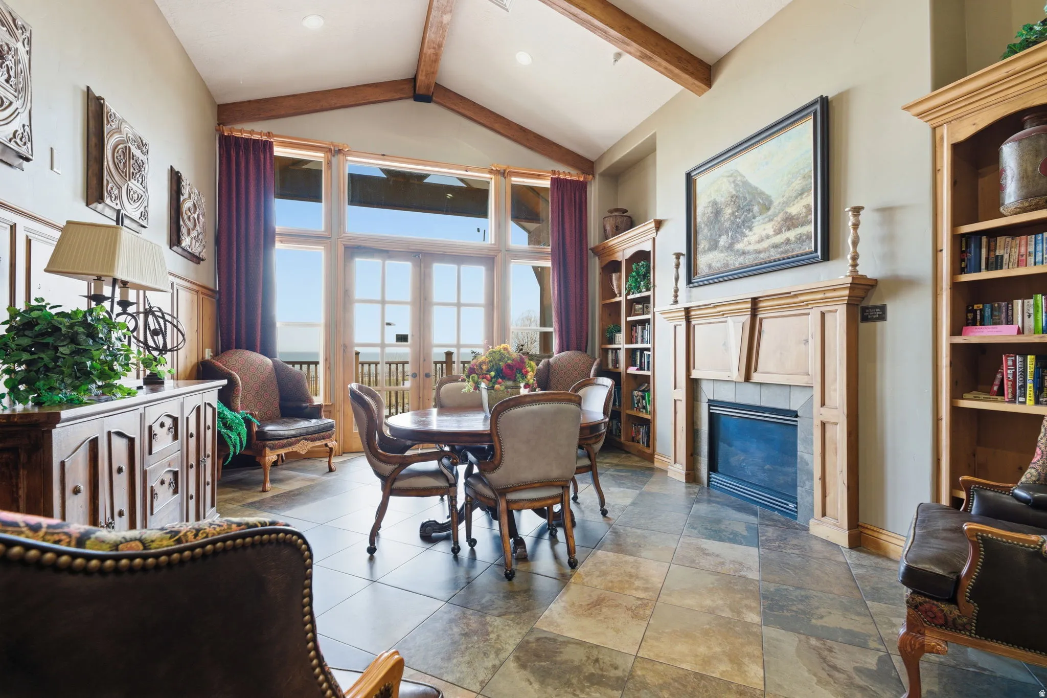 Vaulted ceiling with exposed wood beams, large-format tile flooring, built-in fireplace with wood mantle, built-in shelving, and large glass paneled doors