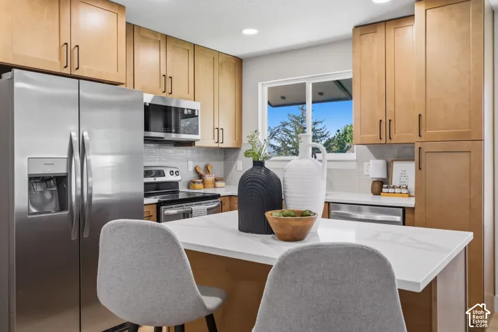 Stainless steel appliances, blonde wood cabinetry, white countertops, tile backsplash, and an island with seating