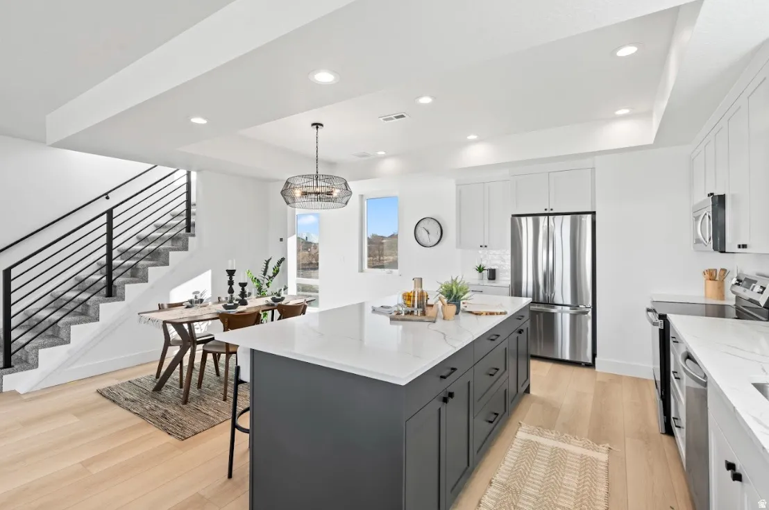 Open-concept living space with light wood-finish flooring, a contemporary kitchen island with a light-colored countertop, white upper cabinetry, and stainless steel appliances