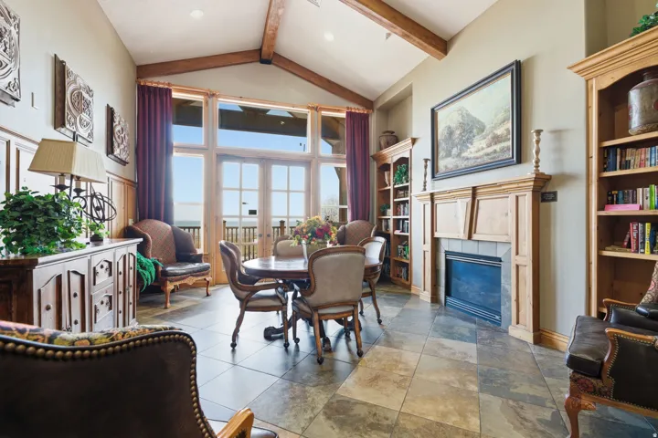 Vaulted ceiling with exposed wood beams, large-format tile flooring, built-in fireplace with wood mantle, built-in shelving, and large glass paneled doors