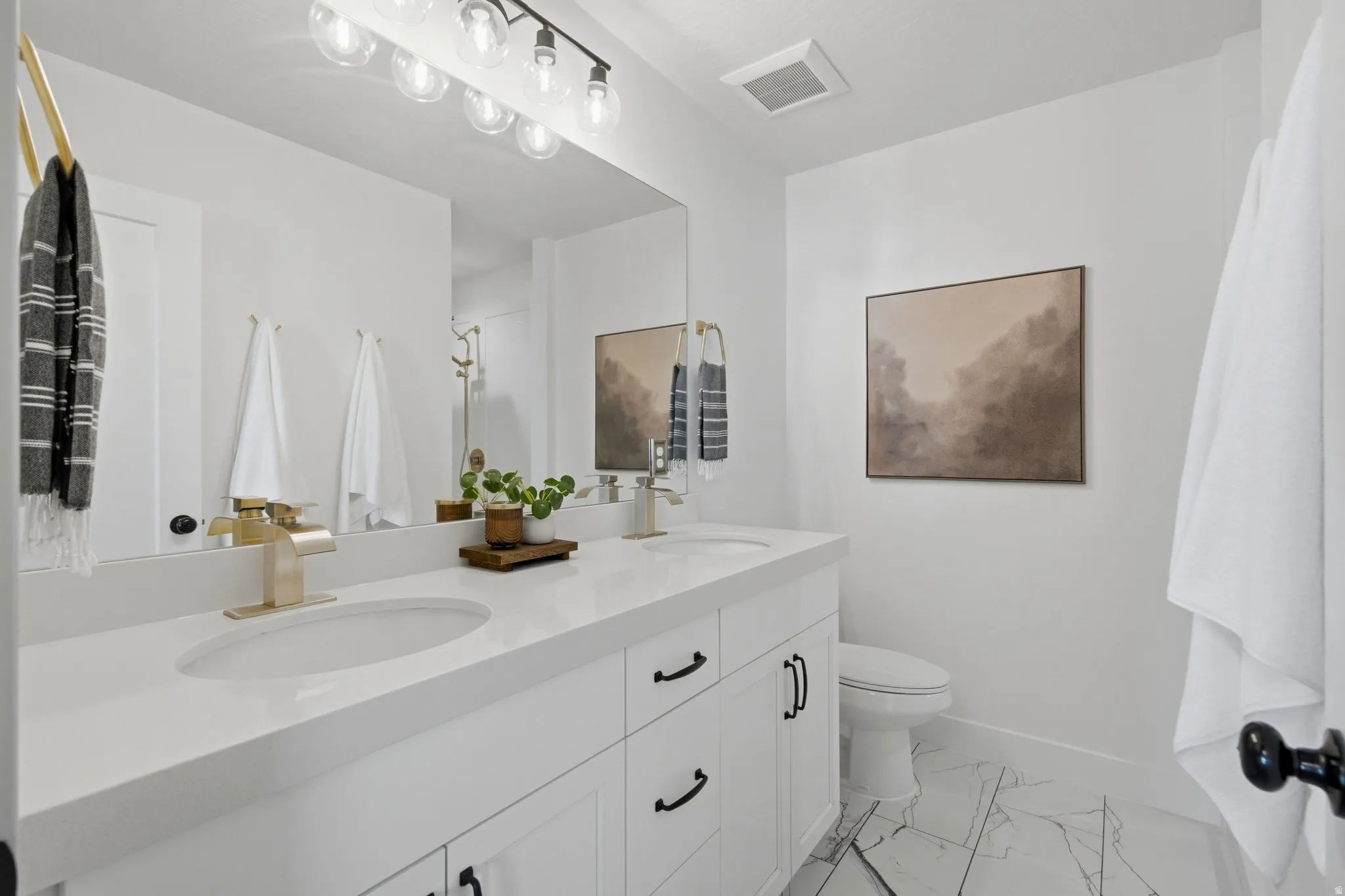 Dual vanity with white cabinetry and black hardware, white countertop, individual sinks with brass-finish fixtures, large vanity mirror, and patterned tile flooring