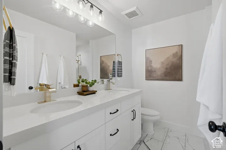 Dual vanity with white cabinetry and black hardware, white countertop, individual sinks with brass-finish fixtures, large vanity mirror, and patterned tile flooring