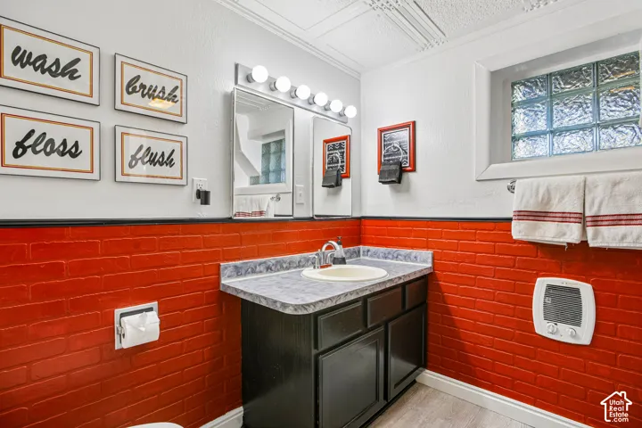 Vanity with integrated sink and dark wood-finish cabinetry, bright wall-mounted light fixture, glass block window, and red brick-style wainscoting