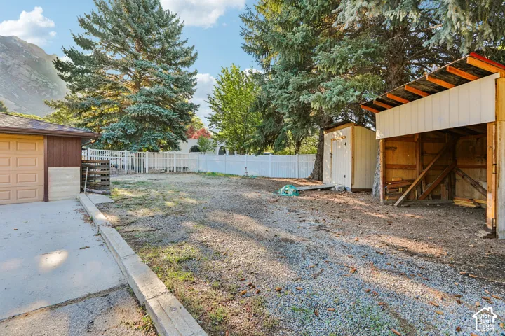 Garage structure with a paneled overhead door, gravel driveway, white privacy fencing, and two storage sheds