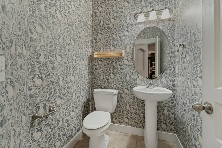 Pedestal sink with chrome faucet, oval mirror, wall-mounted vanity lighting, and patterned wallpaper