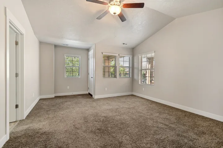 Vaulted ceiling, ceiling fan with integrated lighting, multiple windows with blinds, neutral wall tones, and carpet flooring