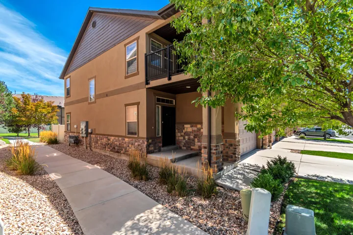 Stucco exterior with stone accent detailing, second-story balcony with black railings, covered entryway, attached garage with overhead door, and concrete walkway