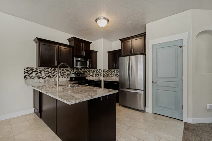 Kitchen featuring an island with a light-toned countertop, dark wood-finish cabinetry, stainless steel appliances, a mosaic tile backsplash, and light-toned tile flooring