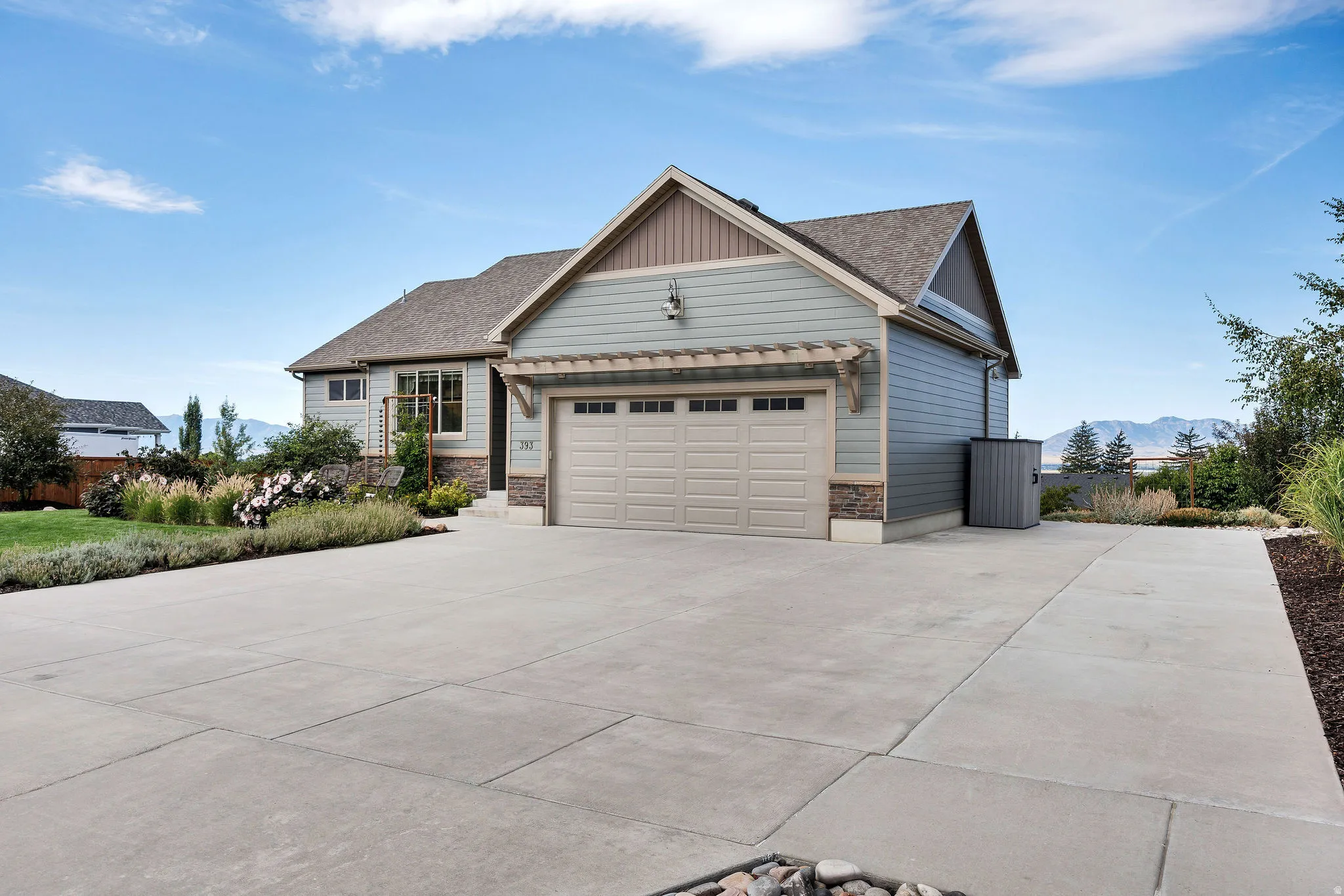 Expansive concrete driveway, attached two-car garage with overhead door, landscaped front yard with shrubs and flowering plants, and mountain scenery in the background