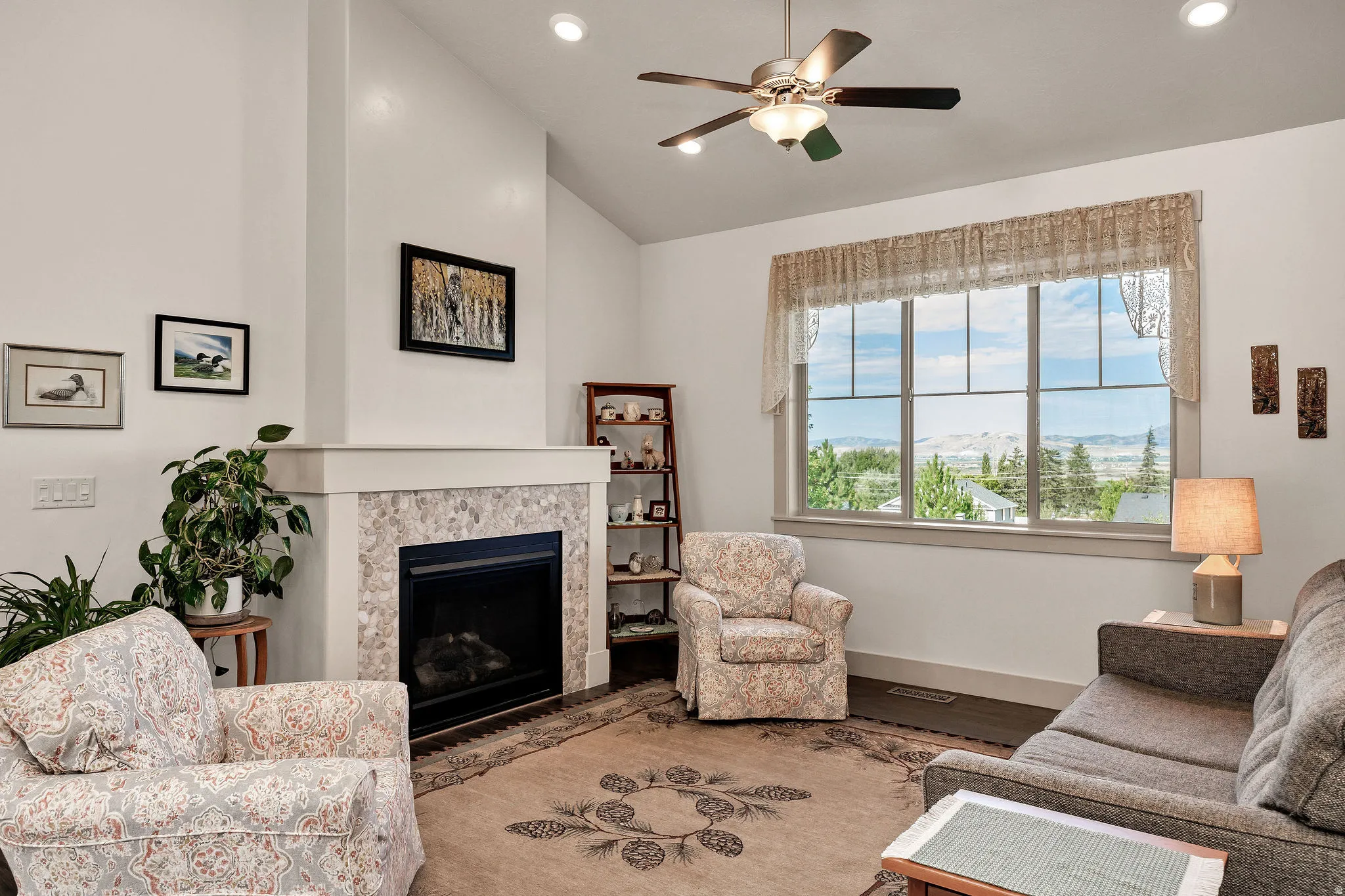 Vaulted ceiling with recessed lighting, ceiling fan with integrated lighting, tiled fireplace, built-in shelving, and large window with mountain scenery