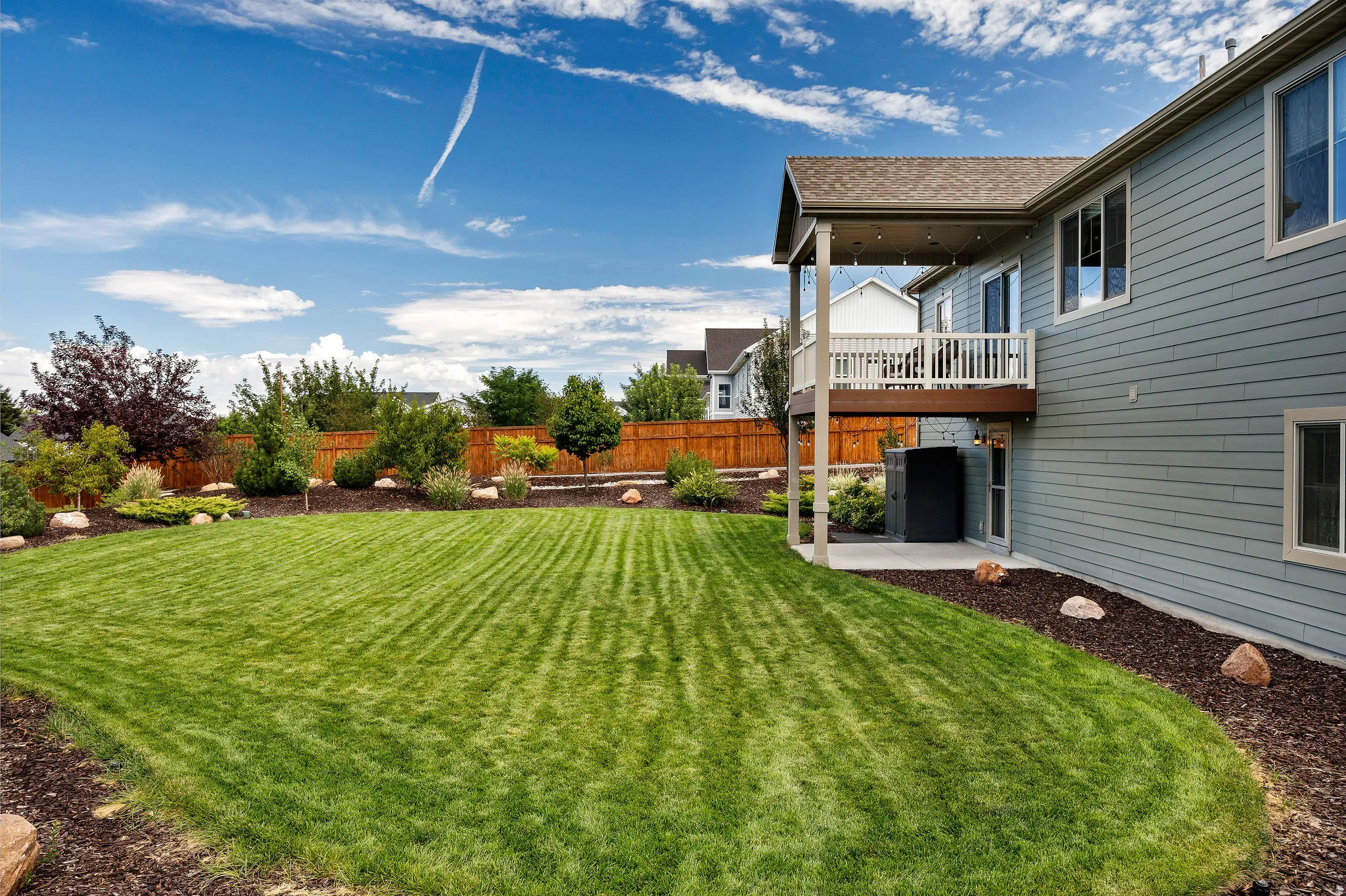 Expansive lawn with perimeter landscaping, sheltered patio, elevated deck with a white railing, and a privacy fence