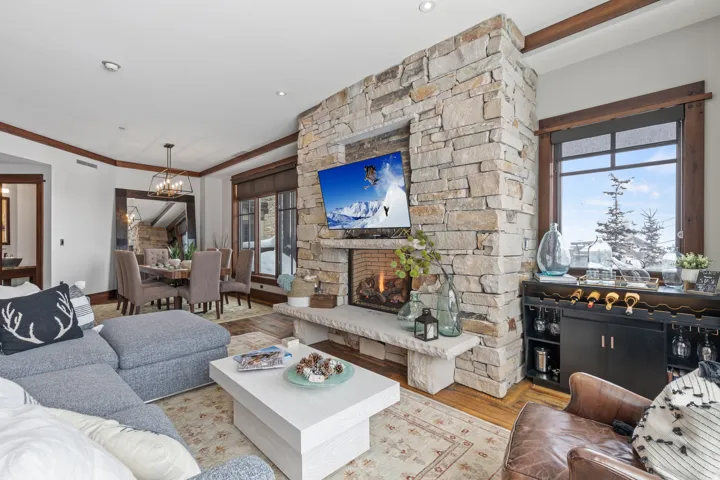 Open-concept living area featuring a custom stone fireplace with gas insert and flat-screen television mount, hardwood flooring throughout, exposed wood beam ceiling details, and expansive windows framing mountain scenery