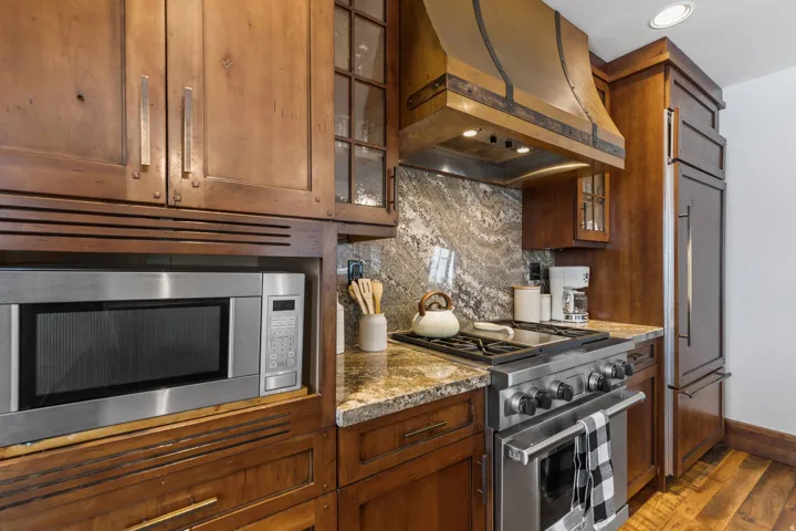 Wood-finish cabinetry with glass accents, stainless steel appliances, granite countertops, and a decorative range hood