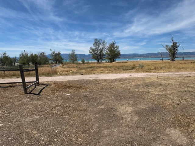 Expansive outdoor space featuring a wooden fence, dirt and dry grass ground cover, mature trees, and a distant body of water