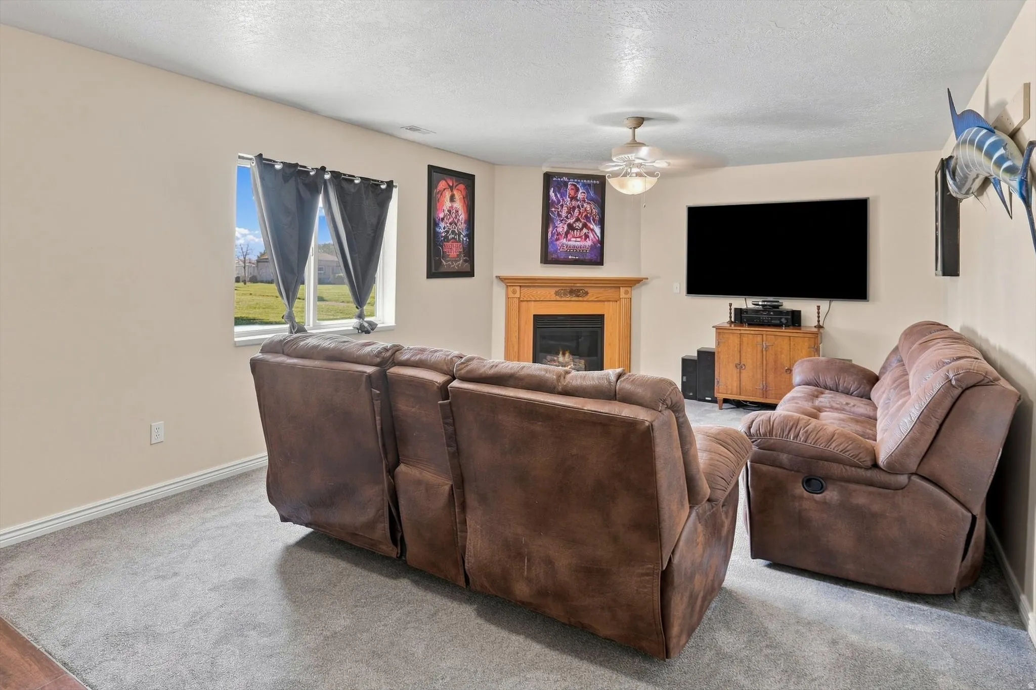 Living area featuring plush gray carpeting, a window with exterior views, a ceiling fan, and a fireplace with a wood mantel