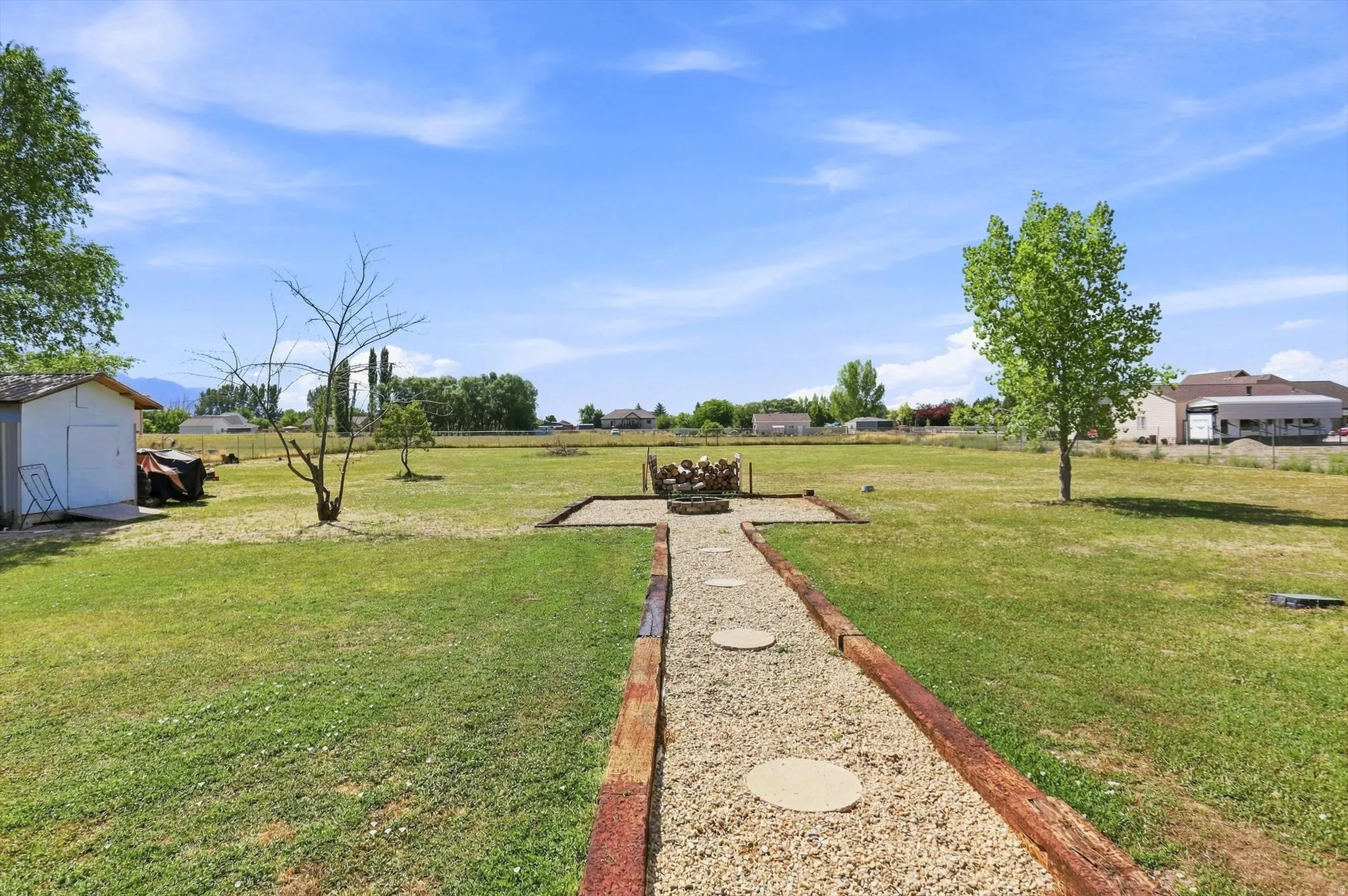 Expansive grassy yard featuring a gravel pathway with stepping stones, a built-in fire pit, and mature trees
