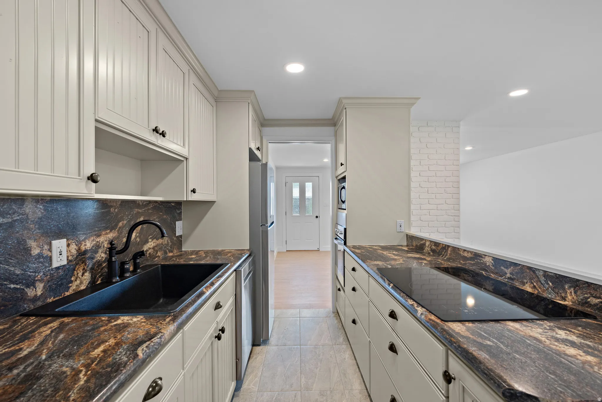 Galley kitchen featuring light-toned cabinetry, dark patterned countertops, a built-in cooktop, and an undermount sink with a matte black faucet