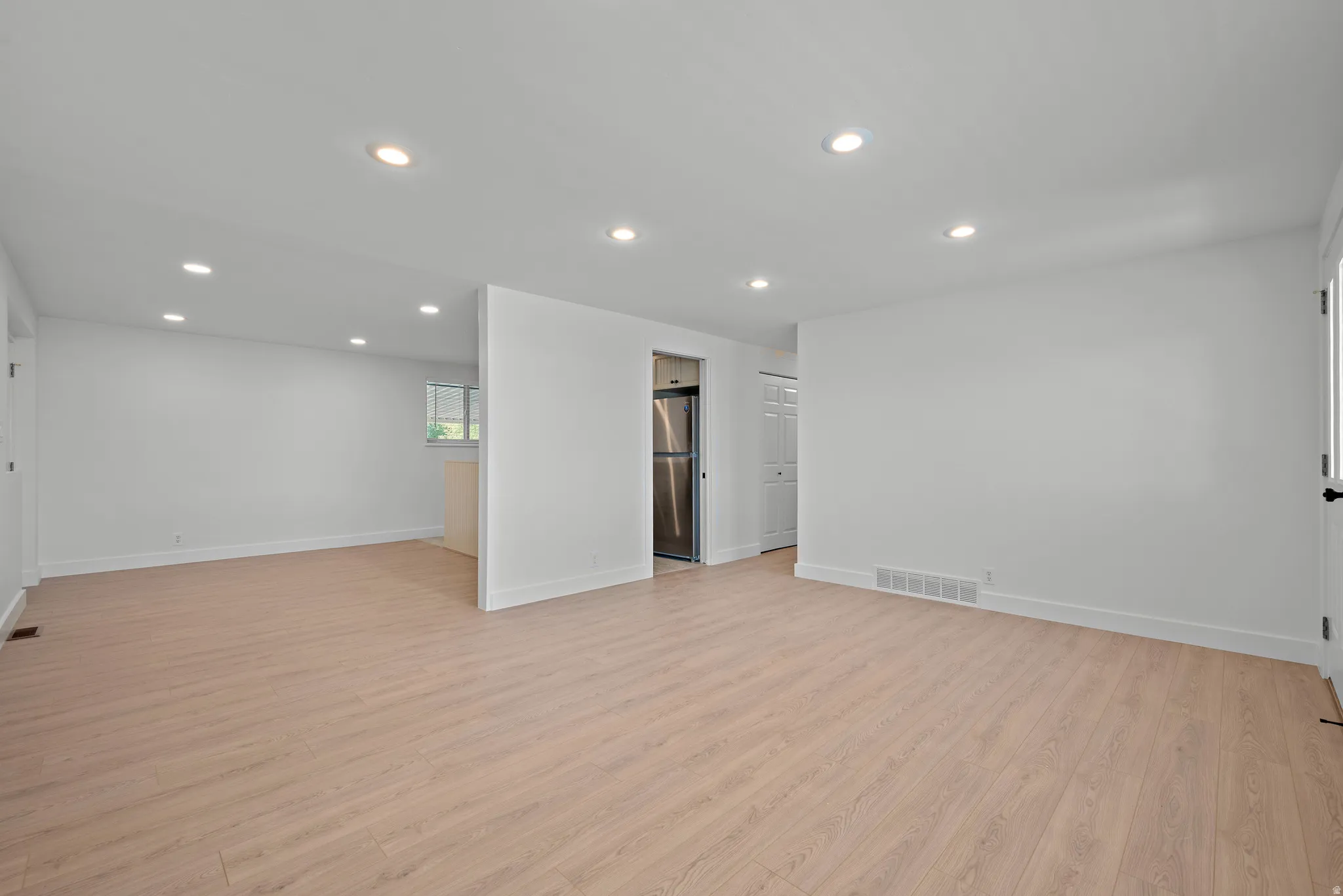 Recessed ceiling lighting, light wood-finish flooring, white painted walls, and a stainless steel refrigerator