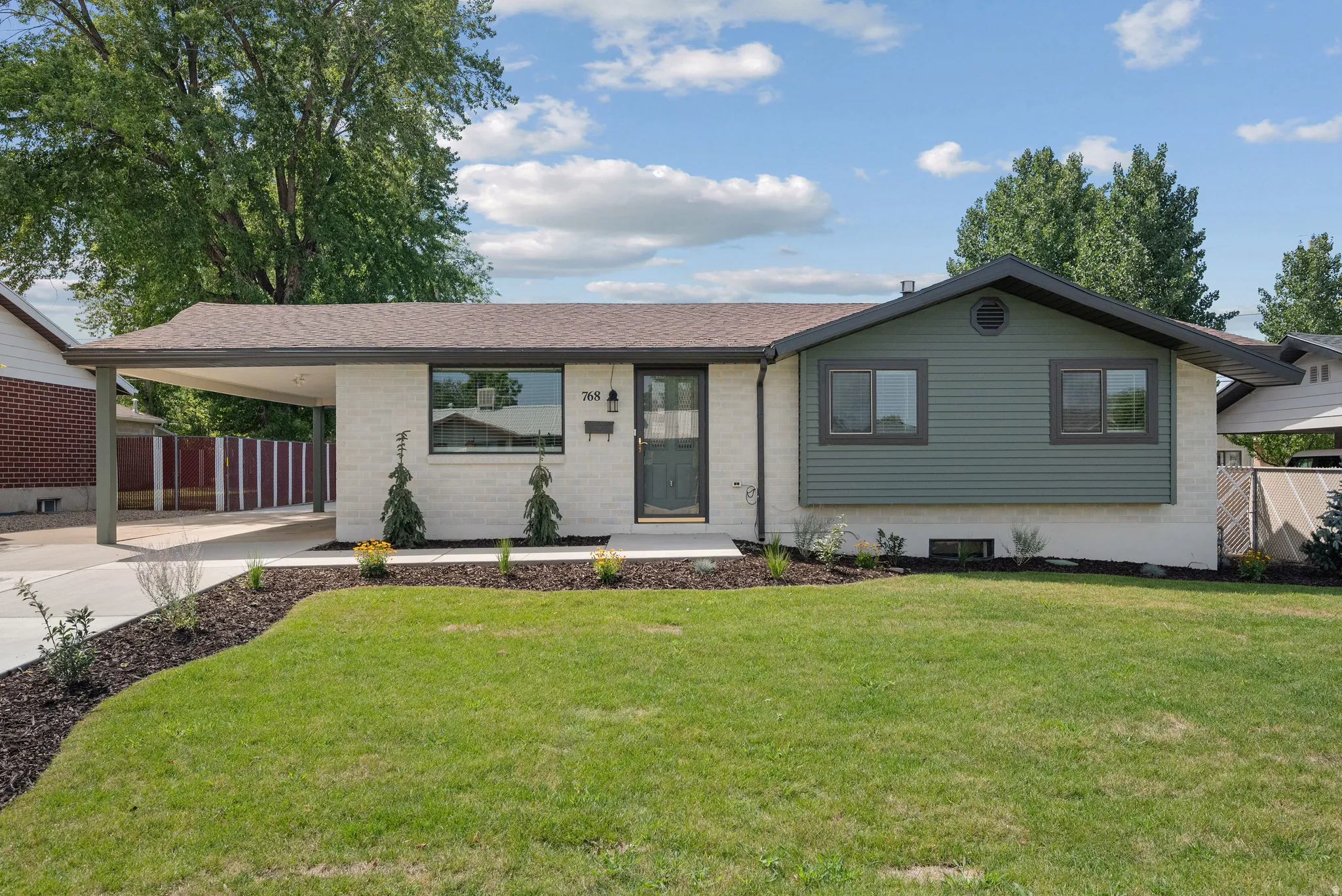 Front exterior with a carport, white painted brick, green siding, a dark roof, and mature landscaping